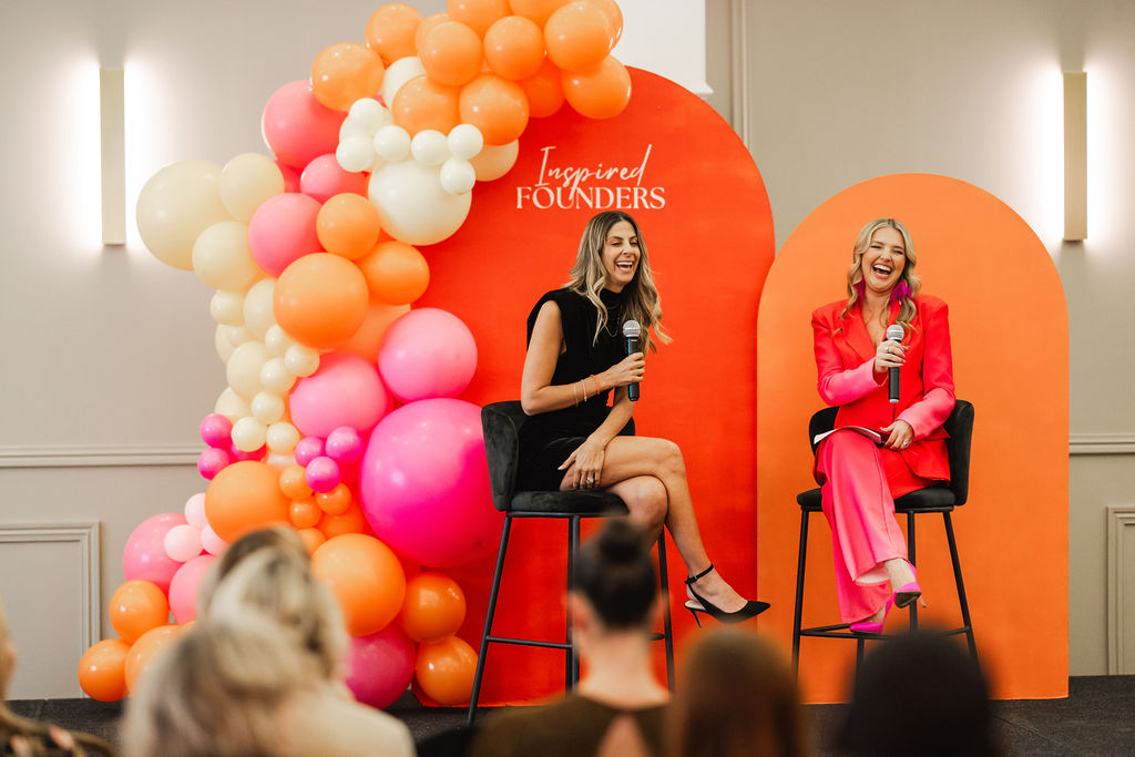 Two speakers laughing together on stage in front of a balloon arch
